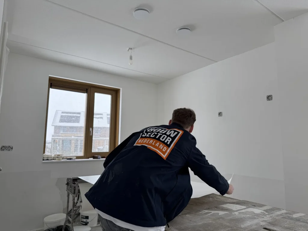 Vakman in bedrijfsjas pakt een baan vlies van de plaktafel in een lege kamer